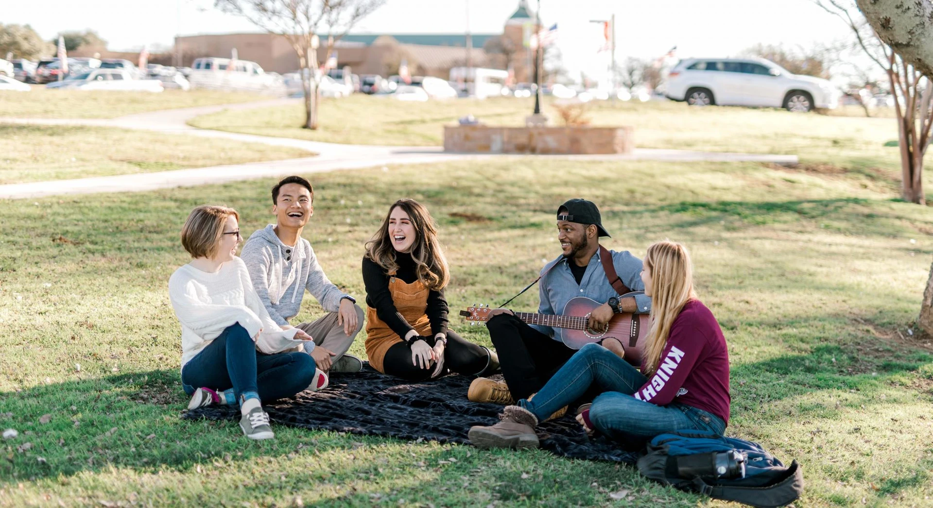 Five students sit under a tree on a blanket laughing and singing as one studentplays the guitar