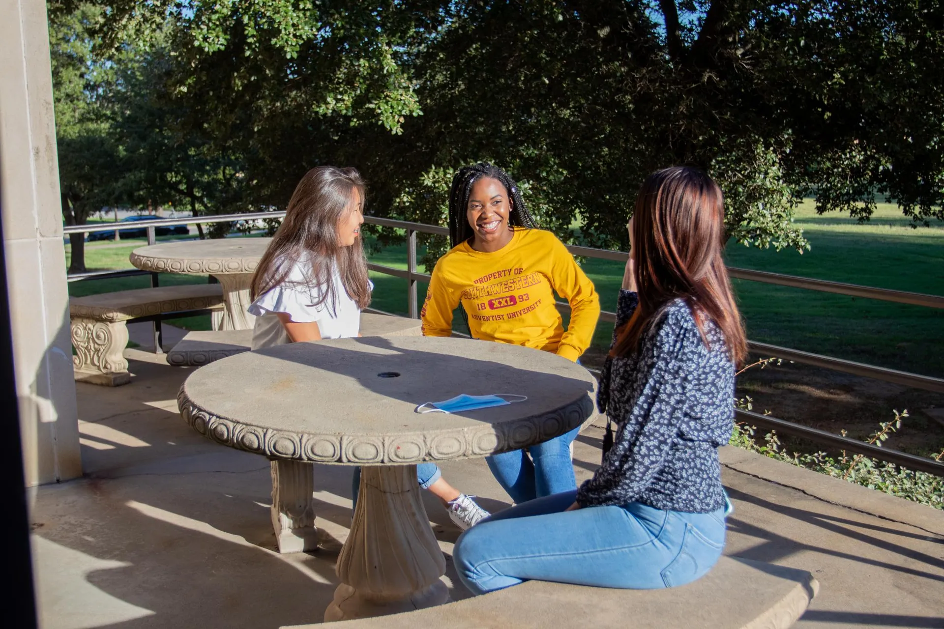 Three female students sit on the patio in the sunny weather and laugh