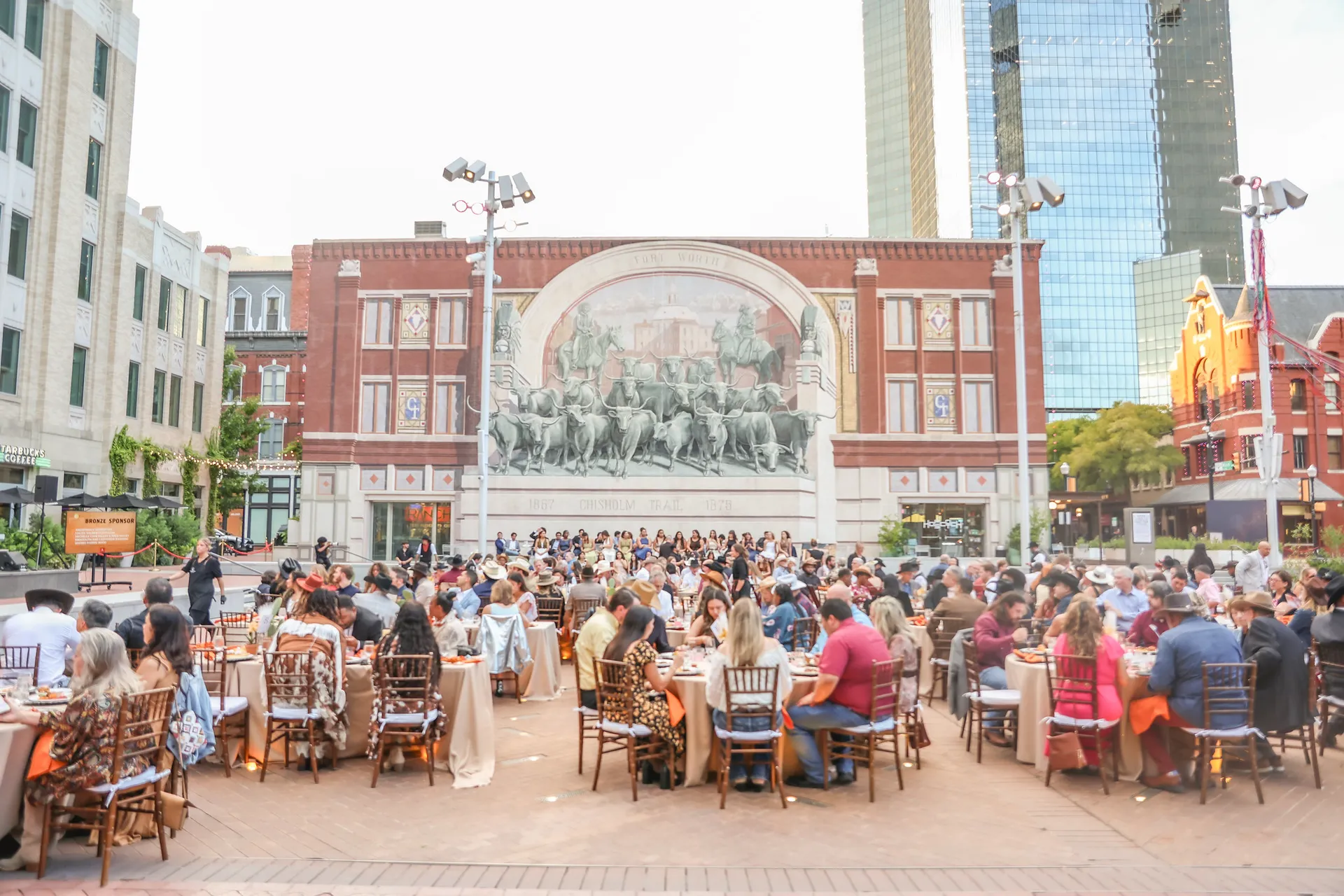 People sitting at dining tables on a patio in front of a large building with a mural