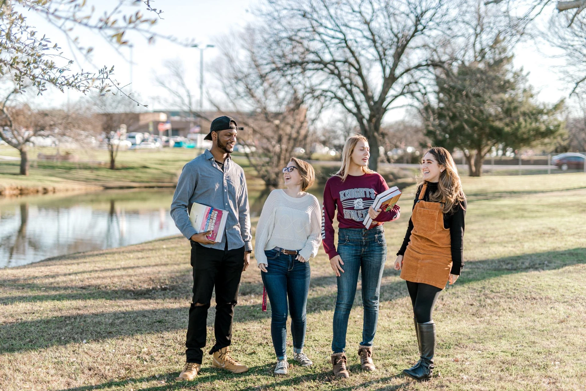 Four students, two with books in hand, laugh as they talk and stand in front of the duck pond.