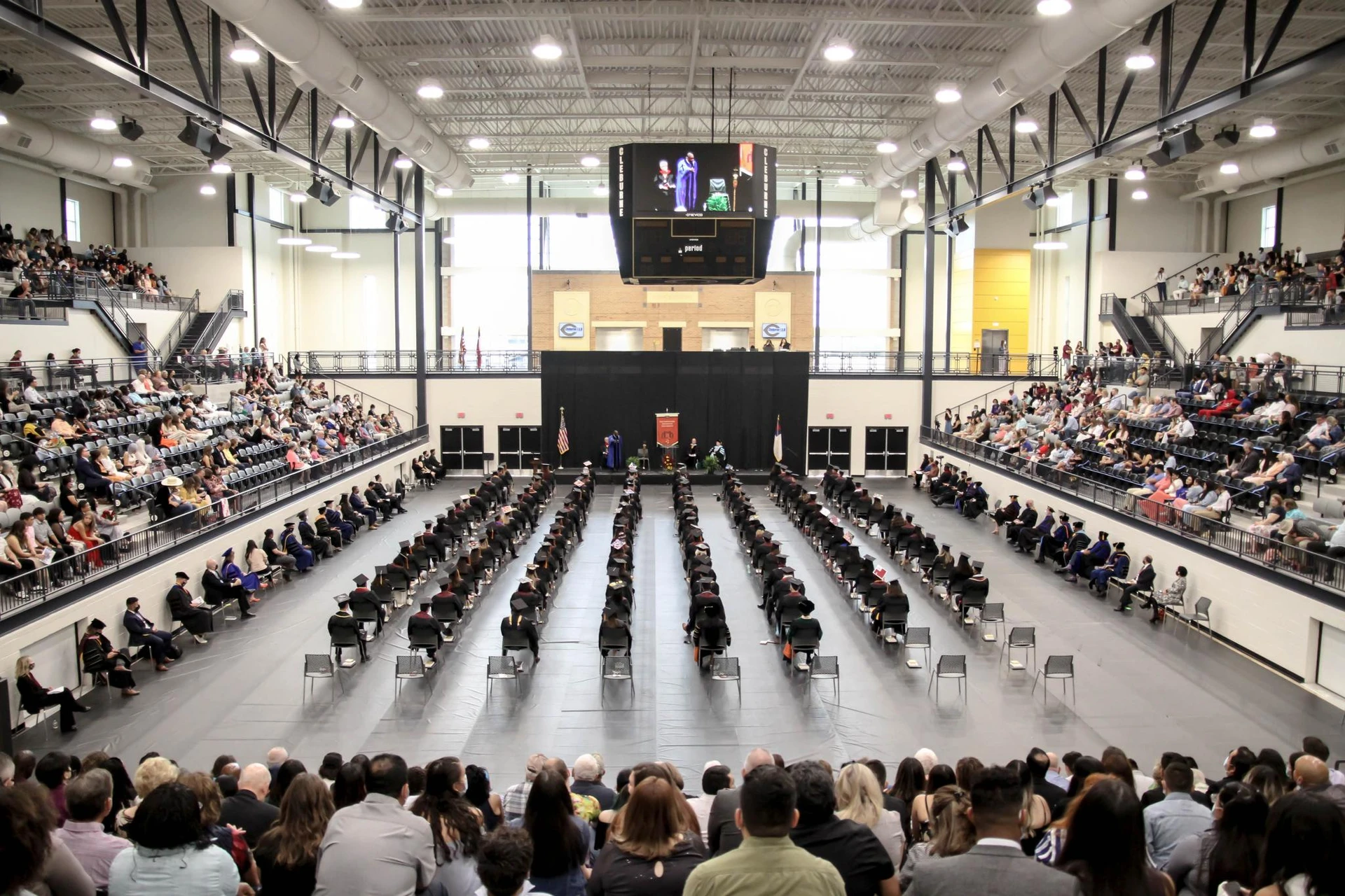 Graduates sitting in rows in a large stadium with guests to the right, left, and back and the stage to the front