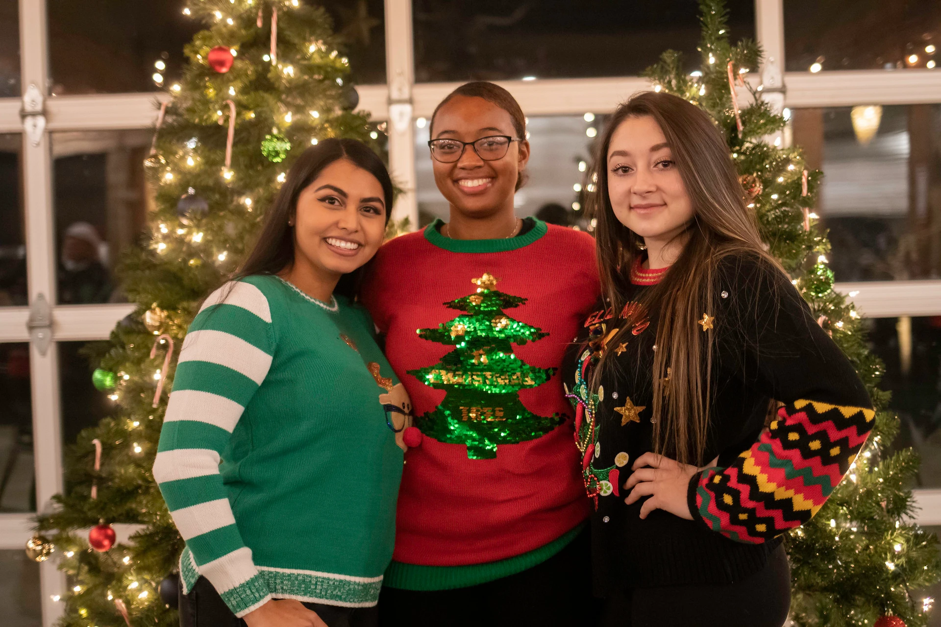 Three young ladies smiling, wearing red and greenn Christmas sweaters, standing together in front of two decorated Christmas trees with their arms around eachother 