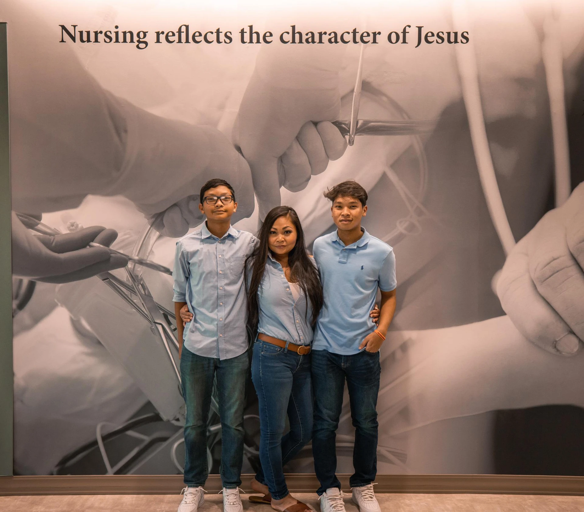 Karen Phongsavan and her two sons pose in front of a wall in the Southwestern Adventist University nursing building that says "Nursing reflects the character of Jesus"