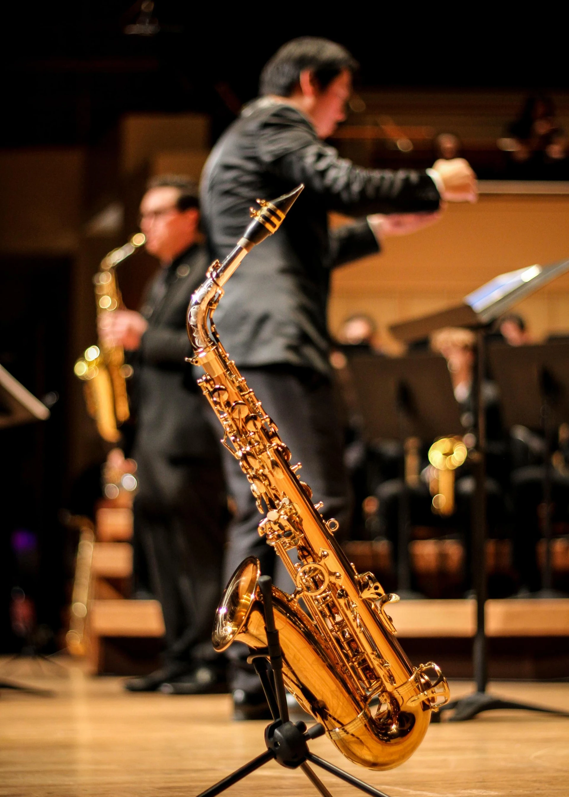 A photo of a shiny saxophone, and in the background a conducter is leading an ensemble