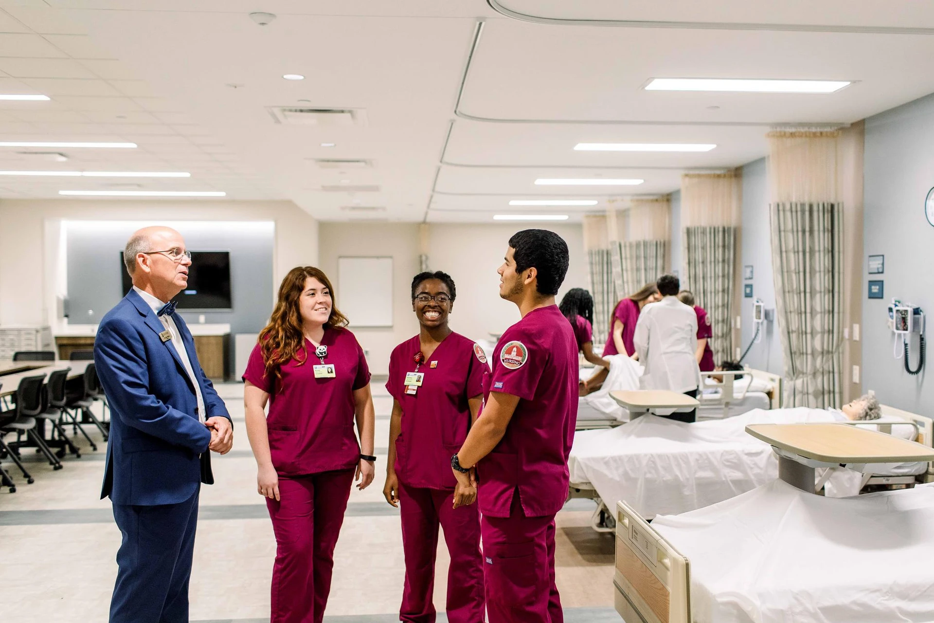 Dr. Shaw, the university president, stand in his blue suit as he speaks with 3 nursing students in scrubs. 