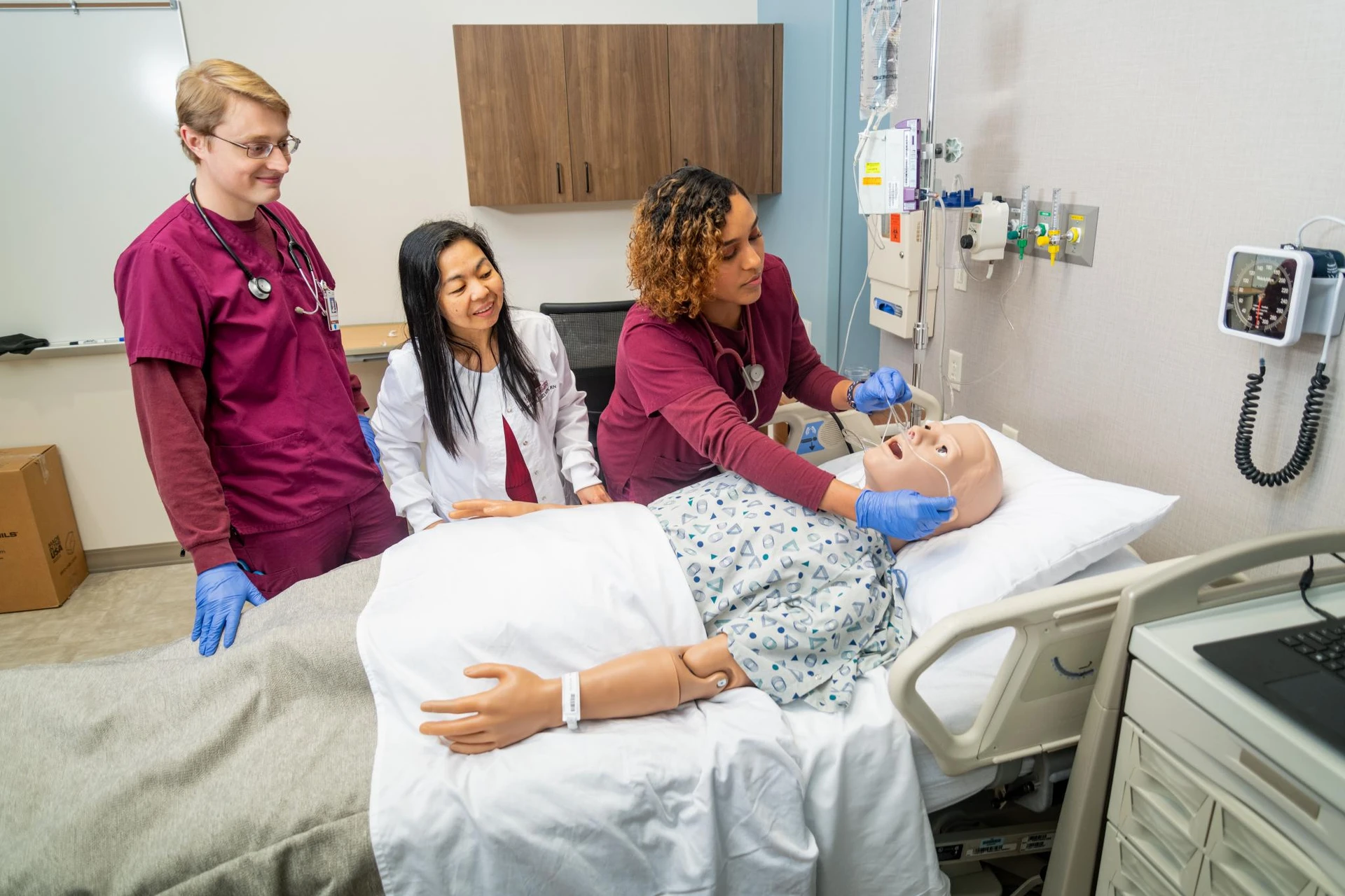 A nursing student, dressed in maroon scrubs, places oxygen tubes in a fake patients nose while a peer and professor stand by and watch