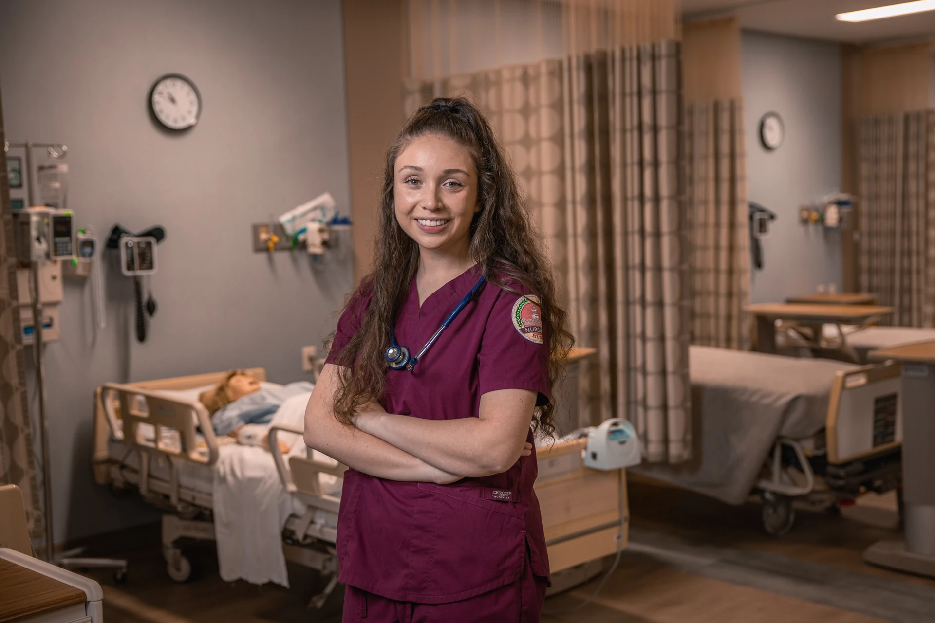 Heather Chirinos poses in the Nursing SIM Lab with a mannequin behind her wearing SWAU scrubs and a stethoscope