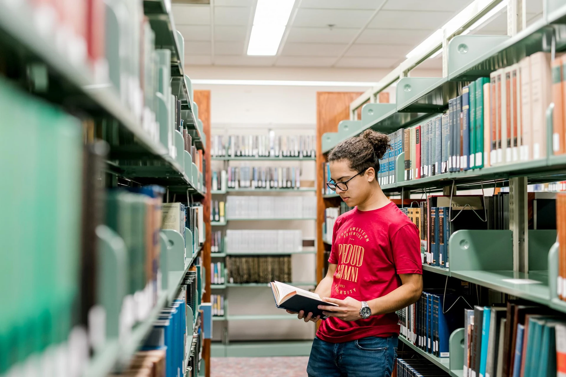 A male student with his hair tied up stands in between the library shelves as he reads through a book